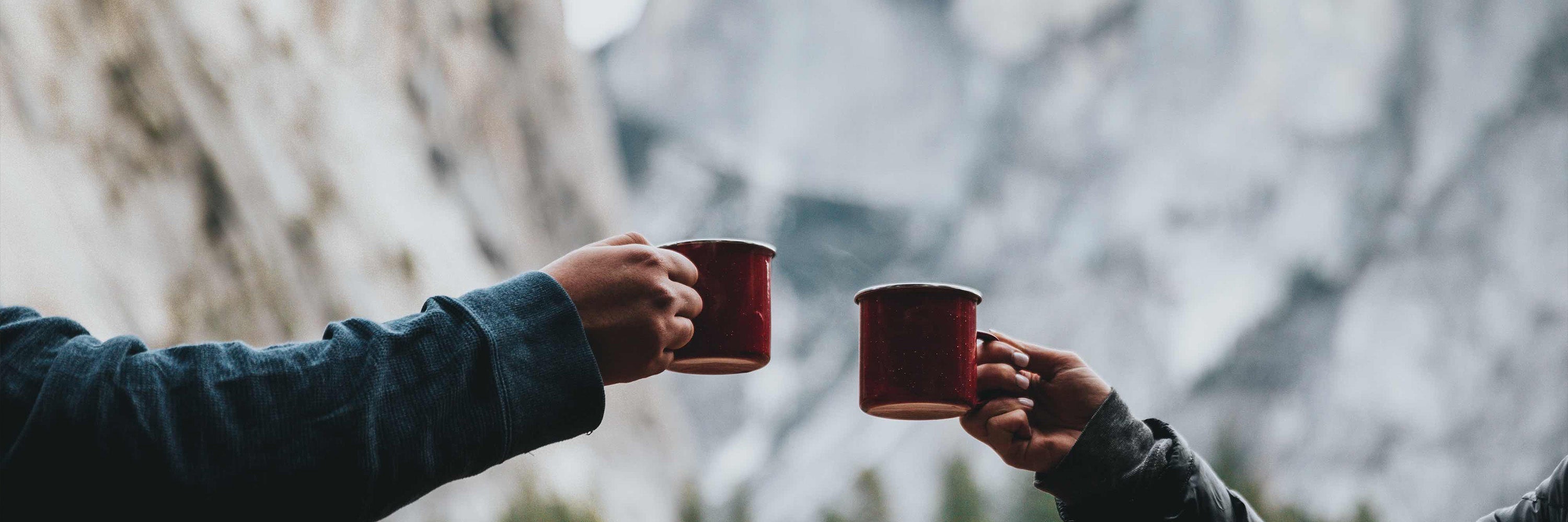 Wide image of two hands holding coffee mugs in front of a mountain range.